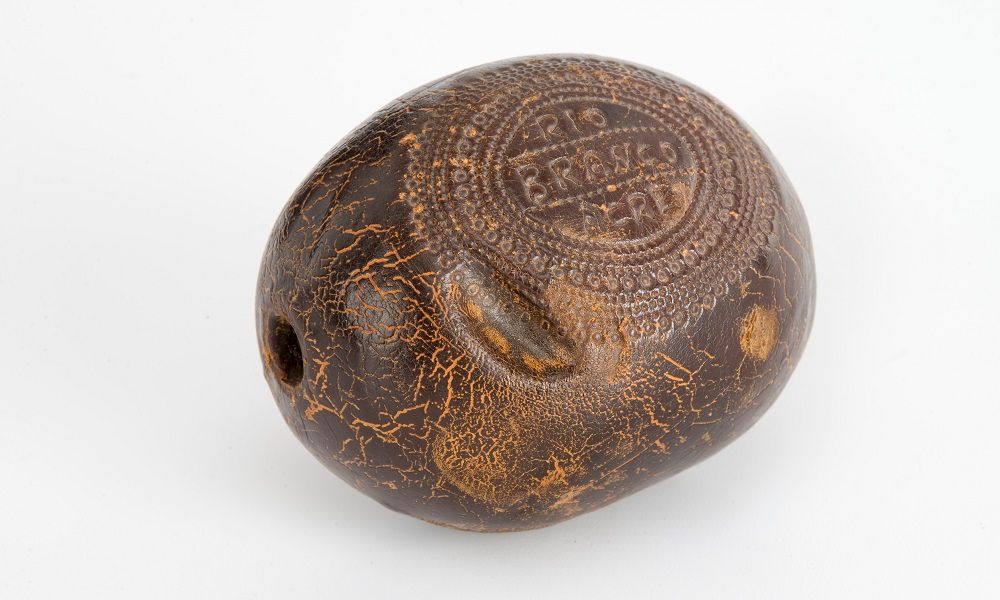 Photographie d'une balle en caoutchouc. L'objet est de couleur marron foncée. Il est possible de lire les inscriptions " Rio Branco Acre". Le texte est entouré d'éléments ornementaux.