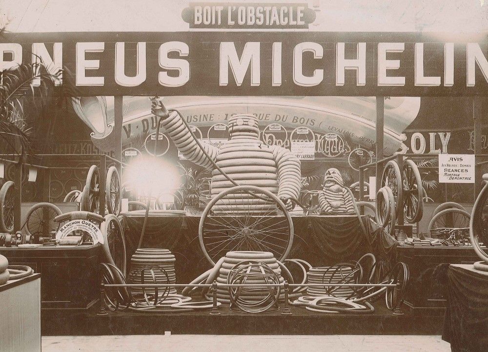Photographie en noir et blanc d'un stand d'exposition Michelin au Salon de l'Automobile de Paris. La scénographie est composée, en son centre, d'une gigantesque sculpture de Bibendum réalisée à partir de pneus. Autour de lui, des pneus de différentes tailles et diamètres sont exposés.