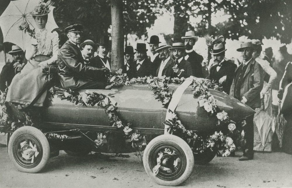 Black and white photograph of a group of individuals gathered around driver Camille Jenatzy, at the wheel of the Jamais Contente. A woman is seated in the back, just behind the driver, in the rear part of the vehicle. The car is decorated with wreaths of flowers and ribbons, symbols of his victory.