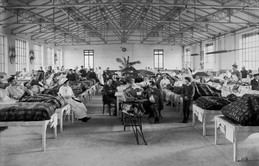 Photographie en noir et blanc montrant un atelier transformé en hôpital de guerre. L'image présente un dortoir où de nombreux soldats blessés sont alités. Des infirmières sont à leur chevet.