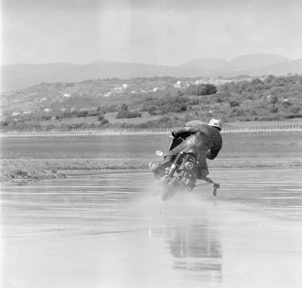 The black-and-white photograph shows a test rider riding a motorcycle on wet ground. His unbalanced posture, leaning to the right, ready to take the turn, is clearly visible.