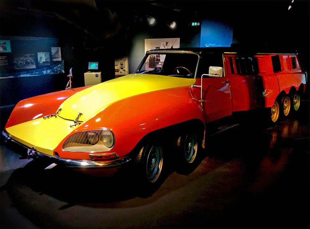 Photograph of the Mille-pattes, an impressive vehicle comparable in length and profile to a limousine. The vehicle is bright red in color, with a wide yellow stripe on the hood. It has a total of 10 wheels, distributed as follows: 4 at the front and 6 at the rear.