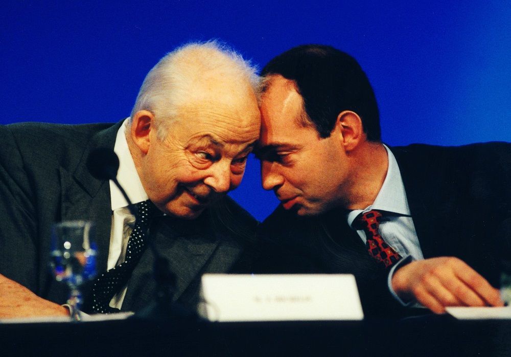 Photograph: close-up portrait of François Michelin (left) and his son Édouard (right). The two men are leaning towards each other, their foreheads so close they are touching. They exchange a knowing glance.