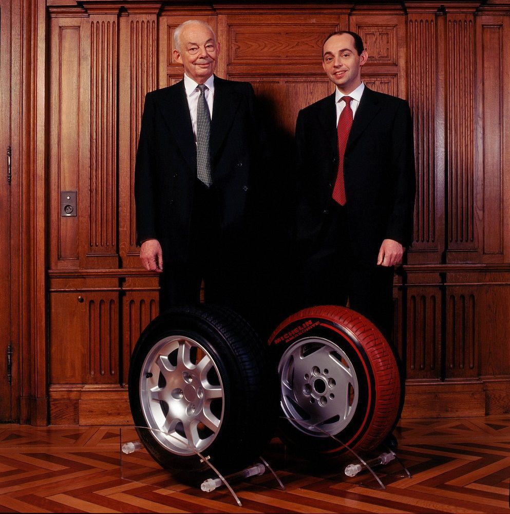 Portrait officiel de François et Édouard Michelin réalisé à l'occasion du centenaire de Bibendum en 1998. La photographie montre les deux hommes côte à côte, souriants. Deux pneus sont disposés devant eux à leurs pieds.