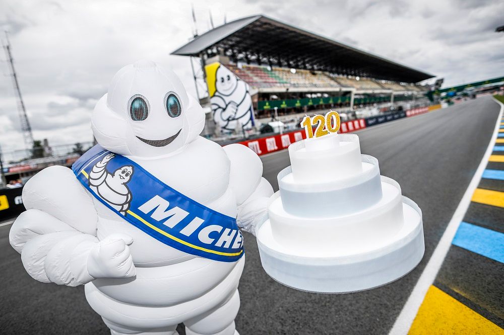 Photographie d'une mascotte de Bibendum sur les pistes de course des 24 Heures du Mans. Il porte un gâteau surmonté de bougies portant le chiffre 120, en référence à son anniversaire.
