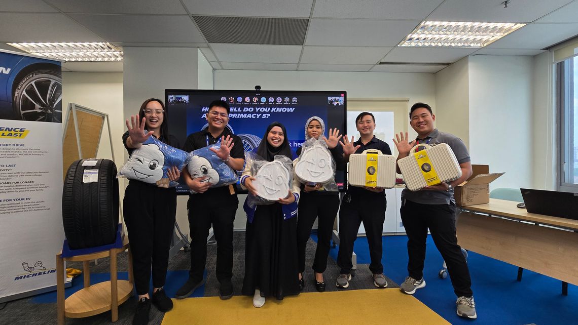 Six employees pose indoors, holding Michelin branded cushions and luggage props, smiling and raising hands.