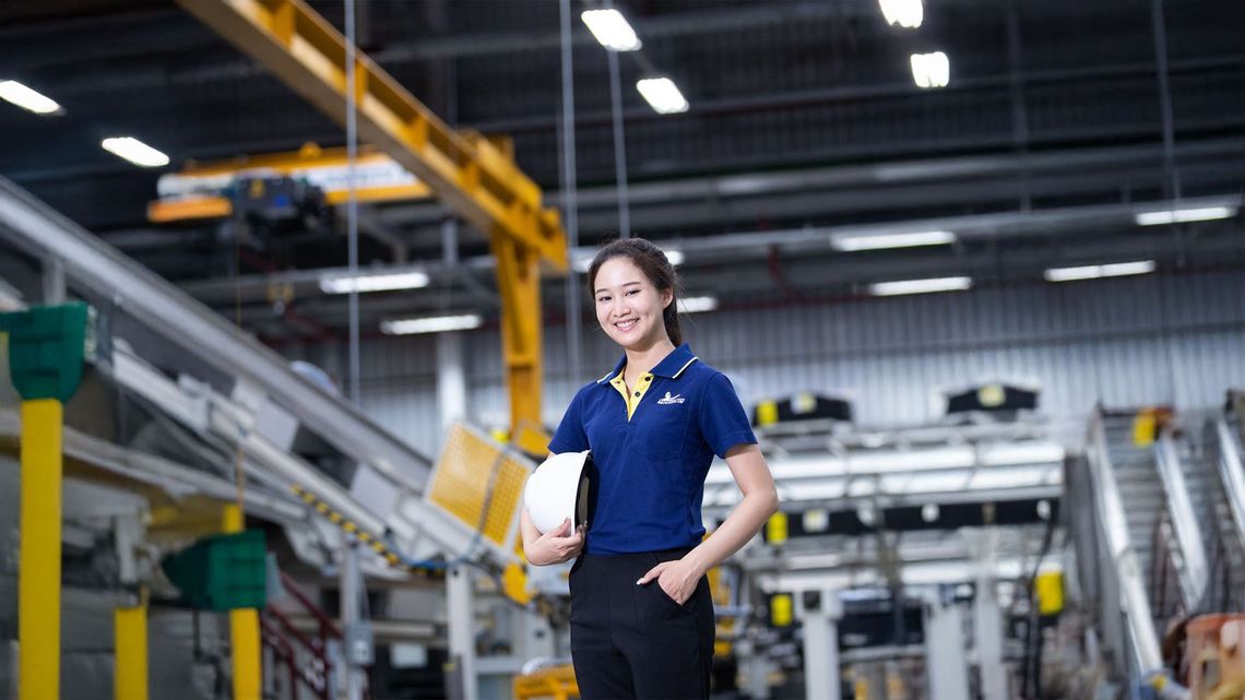 Factory engineer in blue polo holding a white hard hat, standing in an industrial production facility with conveyors and machinery.
